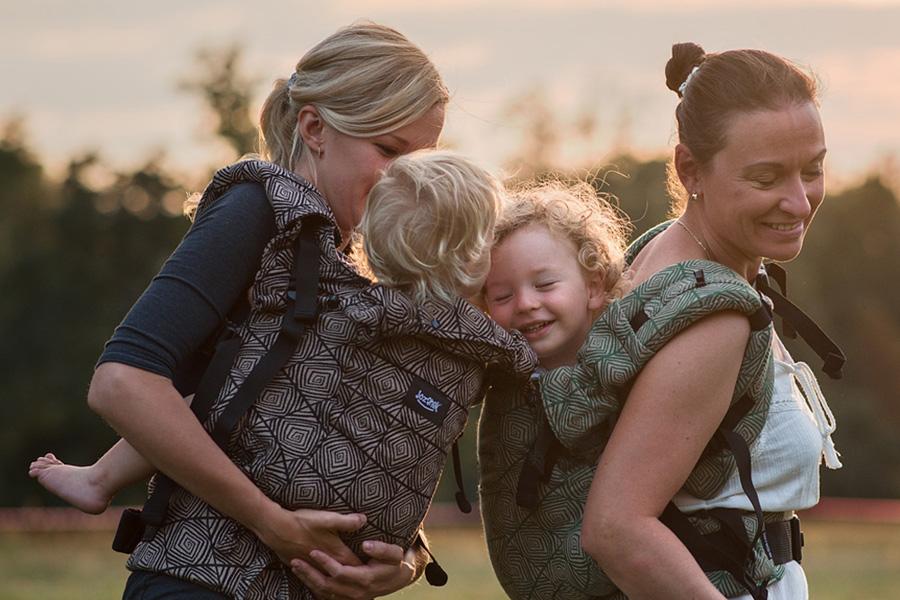 Dve usmievajúce sa matky s batoľatami v nosidlách vpredu na tele